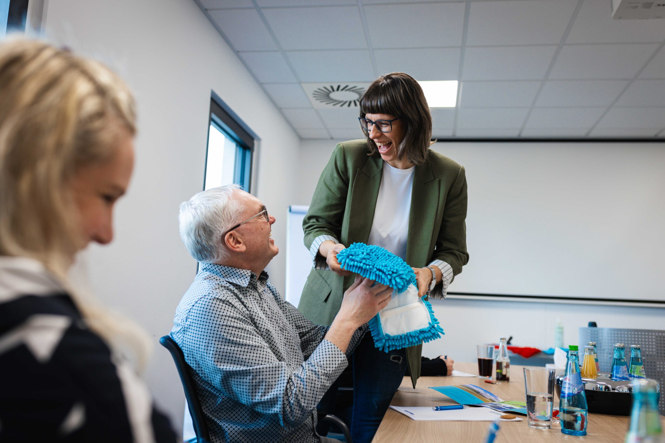 Eine lächelnde Frau im grünen Blazer übergibt einem Mann am Konferenztisch einen blauen Mikrofaser-Moppaufsatz während eines Meetings.